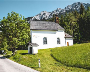 Eine kleine, weiße Kirche steht umgeben von grünen Wiesen und Bäumen. Im Hintergrund sieht man majestätische Berge unter klarem Himmel.