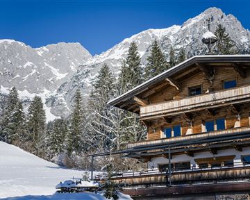 Ein charmantes Chalet im Schnee, umgeben von hohen Bergen und Nadelbäumen. Der Himmel ist klar und blau, was eine winterliche Atmosphäre schafft.