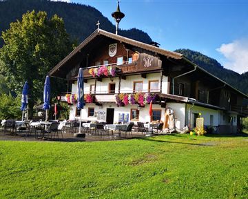 A traditional wooden house with colorful flowers in the windows. It is situated in a green setting with mountains in the background.