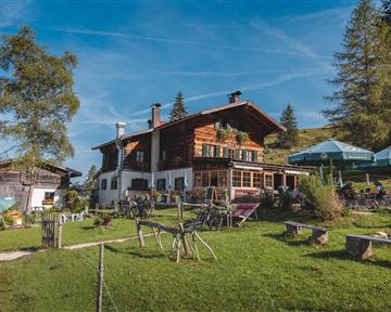 A rustic mountain house surrounded by a green meadow and trees. In the foreground, seating areas and sun umbrellas can be seen.
