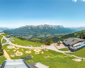 A picturesque mountain landscape with green meadows and snow-covered peaks in the background. In the foreground, playgrounds and buildings can be seen that offer a beautiful view.