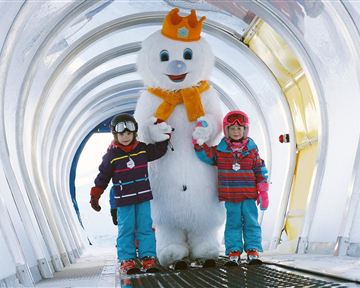 Two children are standing next to a large snowman in a winter alley. They are wearing colorful winter clothes and smiling happily.