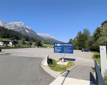 A parking lot in the mountains with a clear view of the surrounding peaks. The sky is blue and the surroundings are green and inviting.