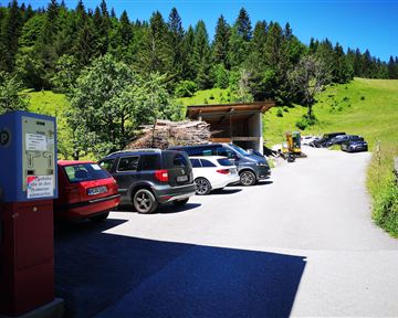 Ein Parkplatz in der Natur, umgeben von grünen Wiesen und Bäumen. Verschiedene Autos sind zu sehen, und im Hintergrund steht eine kleine Scheune.