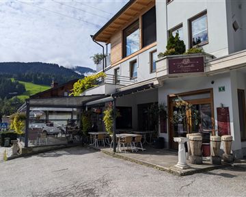 Ein gemütliches Restaurant mit einer Terrasse, umgeben von grünen Bergen. Die Fassade ist modern und einladend, ideal für einen Aufenthalt im Freien.