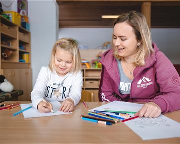A cheerful girl and a young woman are sitting at a table and drawing. Colorful pens and papers are laid out in front of them.