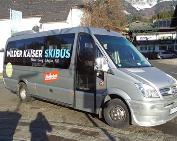 Ein moderner Linienbus für den Skibetrieb, der mit Werbeflächen für den Wilder Kaiser Skibus versehen ist. Der Bus steht auf einem gepflasterten Platz in einer bergigen Umgebung.