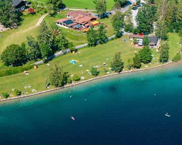 Ein malerischer See mit grünem Gras und Bäumen am Ufer. Im Hintergrund sind einige Gebäude und Menschen, die sich am Wasser entspannen.