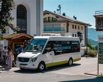 A small bus is driving down a sunny street in a village. In the background, there are houses and some people waiting.