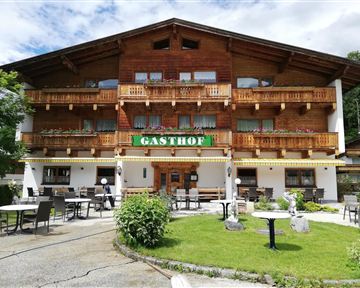A cozy inn with a wooden facade and balconies. The outdoor area features tables and chairs in a green environment.