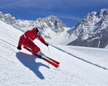 Ein Skifahrer in rot fährt eine schneebedeckte Piste hinunter. Im Hintergrund sind majestätische Berge und ein klarer blauer Himmel zu sehen.