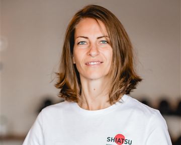 Eine Frau mit braunen Haaren steht lächelnd in einem hellen Raum. Sie trägt ein weißes T-Shirt mit dem Aufdruck "SHIATSU".