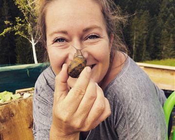 Eine Frau hält einen kleinen Schnecken in der Hand und lächelt in die Kamera. Im Hintergrund sind grüne Bäume und ein Garten sichtbar.