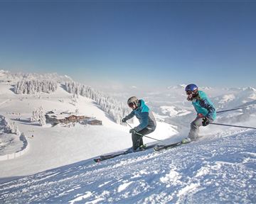 Two skiers on a snow-covered slope with mountains in the background. The sky is clear and the landscape is winterly beautiful.