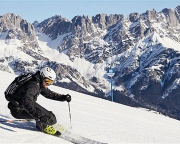 Ein Skifahrer fährt den verschneiten Hang hinunter. Im Hintergrund sind beeindruckende, schneebedeckte Berge zu sehen.