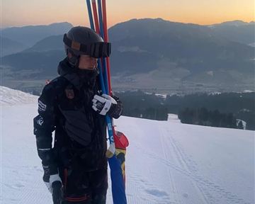 Ein Skifahrer steht auf einer verschneiten Piste mit Skistöcken in der Hand. Im Hintergrund sind die Berge und der Sonnenuntergang zu sehen.