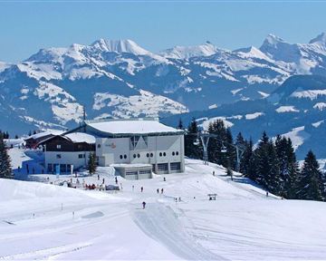 Eine schneebedeckte Berglandschaft mit einer modernen Berghütte. Im Hintergrund sind majestätische Berge und ein klarer blauer Himmel zu sehen.