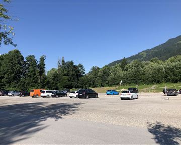 A large parking lot, partially filled, surrounded by trees and mountains. The sky is clear and blue.