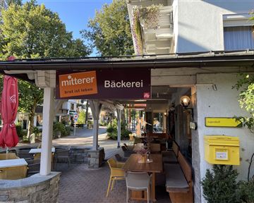 A cozy bakery with an inviting outdoor area. The surroundings are characterized by trees and sunny weather.