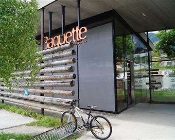 Ein modernes Geschäft mit der Aufschrift „Baguette“. Draußen steht ein Fahrrad und es gibt einen grünen Bereich mit Bäumen.