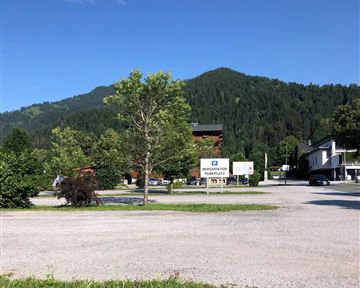 Eine ruhige Landschaft mit grünen Bäumen und einem Blick auf die Berge. Im Vordergrund gibt es einen Platz mit unbefestigtem Boden und Gebäuden im Hintergrund.