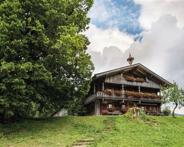 A traditional wooden house in a rural setting, surrounded by trees and green meadows. The sky is partly cloudy and provides an idyllic atmosphere.