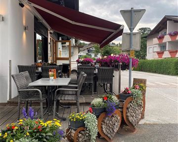 A cozy outdoor area of a restaurant with tables and chairs. Colorful hanging flowers and decorative plants create an inviting atmosphere.