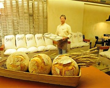 A baker presents fresh bread in a cozy café. In the foreground, there are decoratively arranged loaves and decorative ears of grain.