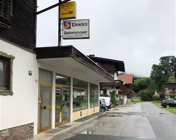 Ein Elektrogeschäft mit dem Namen "Salvenmoser" an einer ruhigen Straße. Im Hintergrund sind typische alpine Gebäude und ein bewölkter Himmel zu sehen.