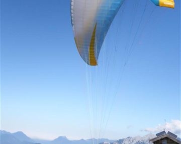 Ein Paraglider startet von einem bewaldeten Hang. Im Hintergrund sind Berge und ein klarer Himmel zu sehen.