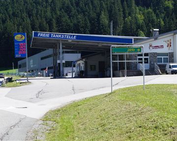 Eine Tankstelle mit dem Schild "Freie Tankstelle" in einer grünen Berglandschaft. Im Vordergrund sind Autos und Wegweiser zu sehen.