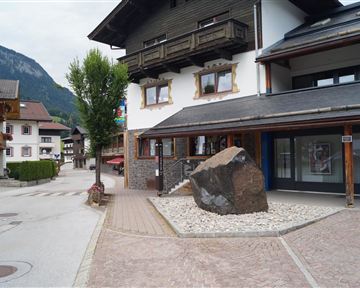 A quiet street with traditional buildings and a large stone in the middle. In the background, green mountains are visible.