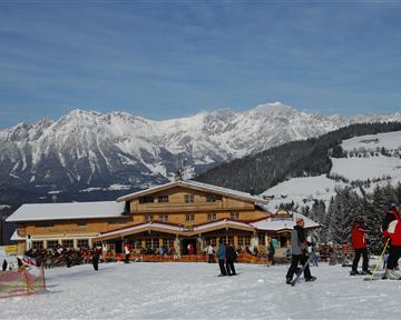 Eine alpine Skilandschaft mit einem gemütlichen Chalet und schneebedeckten Bergen im Hintergrund. Skifahrer und Besucher genießen das Winterwetter.