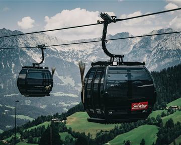 Eine Seilbahn fährt durch eine malerische Berglandschaft. Im Hintergrund sind hohe Berge und grüne Wiesen zu sehen.