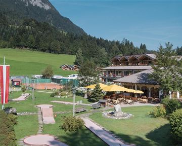 A picturesque hotel in the mountains with a beautiful garden and a mini-golf course. The clear sky and the green landscape create a relaxed atmosphere.