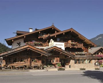 Ein traditionelles Alpenhaus mit Holzfassade und Balkonen. Im Vordergrund befindet sich eine Terrasse mit Sitzgelegenheiten und blühenden Blumen.