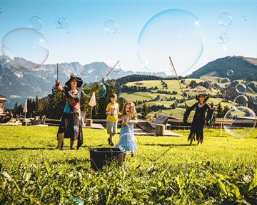 Ein Mädchen beugt sich über eine Wasserrinne und beobachtet das fließende Wasser. Im Hintergrund steht ein Junge und schaut neugierig zu.