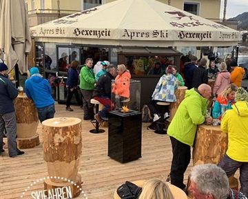 A lively après-ski area with many people gathered around wooden logs and a grill. In the background, a chalet with a large sun umbrella can be seen.