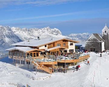 Ein schneebedecktes Bergdorf mit einem modernen Restaurant und einer alten Kirche. Die Umgebung ist von majestätischen Bergen und klarem Himmel umgeben.