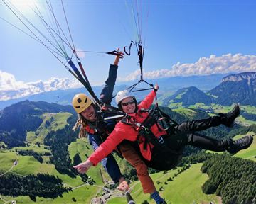 Zwei Personen beim Paragliden über einer grünen, hügeligen Landschaft. Der Himmel ist blau mit einigen Wolken.