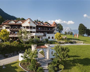 A picturesque hotel in alpine style, surrounded by green meadows and trees. Majestic mountains can be seen in the background.