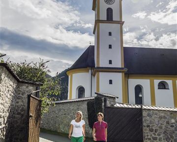 Zwei Personen gehen auf einem Weg vor einer Kirche. Der Himmel ist klar und die Sonne scheint.