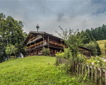 Ein traditionelles Holzhaus umgeben von grünen Wiesen und Bäumen. Im Hintergrund sind sanfte Hügel und eine bewölkte Himmel zu sehen.