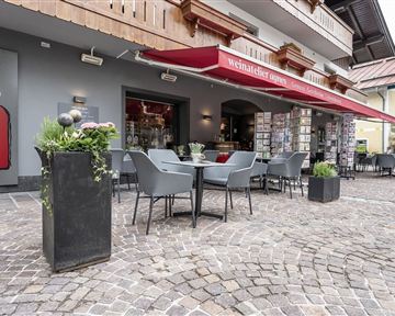 Ein gemütlicher Außenbereich eines Cafés mit Tischen und Stühlen. Rote Sonnenschirme bieten Schatten, und dekorative Pflanzen sind ebenfalls vorhanden.