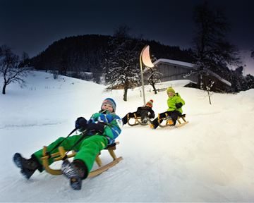 Drei Kinder rodeln fröhlich auf einem verschneiten Hang. Im Hintergrund ist ein Holzhaus und Bäume zu sehen.