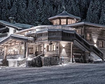 A cozy wooden house in the snow, surrounded by tall fir trees. The surroundings are wintry and peaceful.