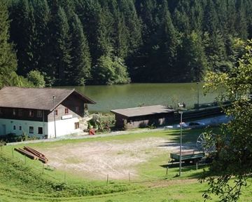 Eine malerische Landschaft mit einem ruhigen See und umgebenden Wäldern. Im Vordergrund sind mehrere Gebäude und ein Platz zu sehen.