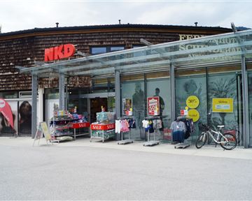 Ein Geschäft von NKD mit einer modernen Glasfassade. Vor dem Laden stehen Kleidung und Werbetafeln mit Angeboten.