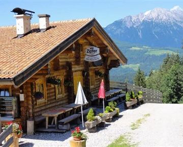 A traditional mountain cabin constructed of wood with an inviting outdoor area. In the background, majestic mountains and a clear blue sky can be seen.