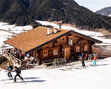 A cozy wooden house in the mountains, surrounded by snow-covered slopes. Skiers enjoy the piste in front of the cabin.